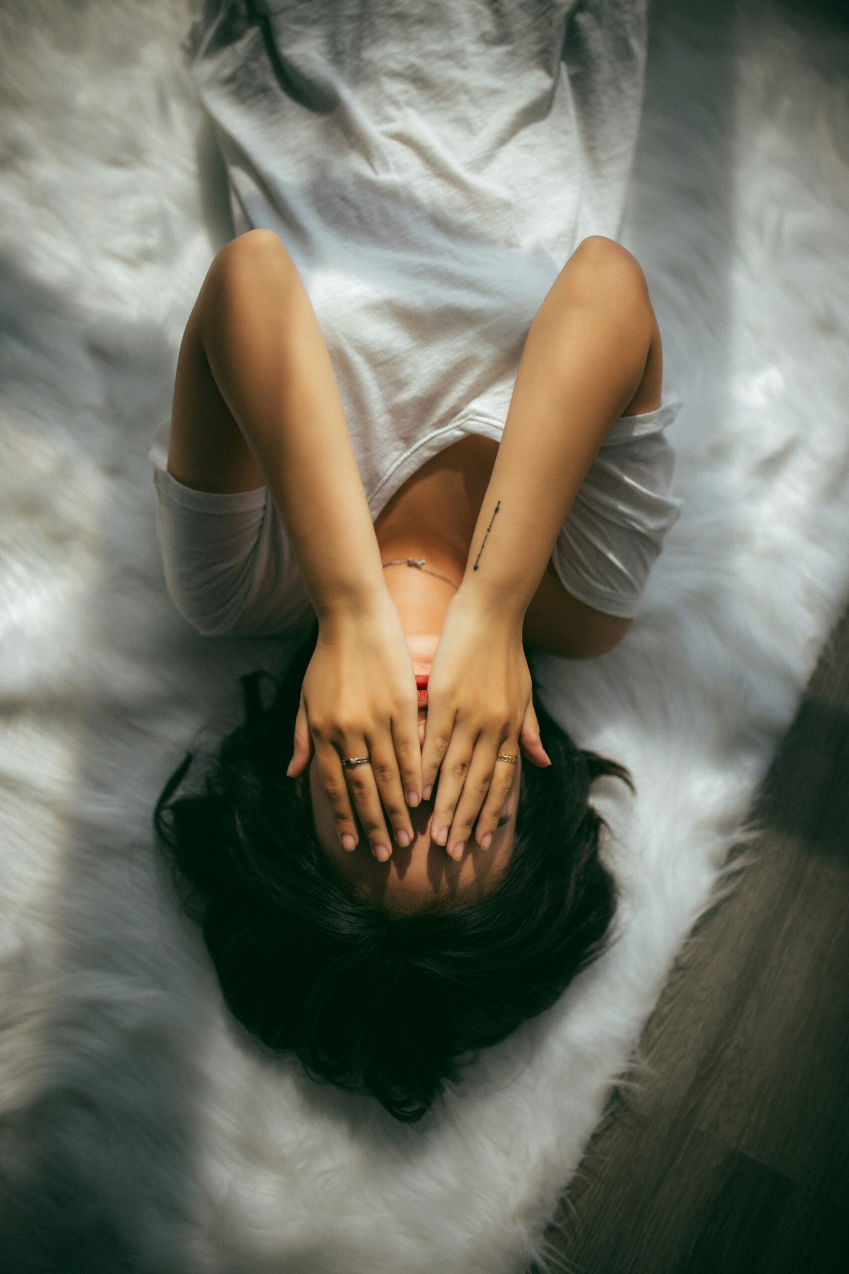 Une personne allongée sur le dos, les mains sur le visage, dans une posture de retrait ou d’épuisement. Vêtue d’un t-shirt blanc simple, elle semble chercher un refuge dans le calme d’un espace doux et neutre. Cette image évoque la fatigue mentale, l’isolement et le besoin de pause des signes fréquents du burn-out. Le regard caché, les jambes repliées : un corps qui se ferme, comme pour se protéger. **Des solutions existent** : un accompagnement bienveillant et personnalisé est proposé à l’Espace des Murmures, pour retrouver l’équilibre, la confiance et la sérénité.